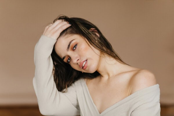 A person with long brown hair and light skin touches their head, wearing an off-shoulder cream top, gazing softly at the camera against a neutral background.