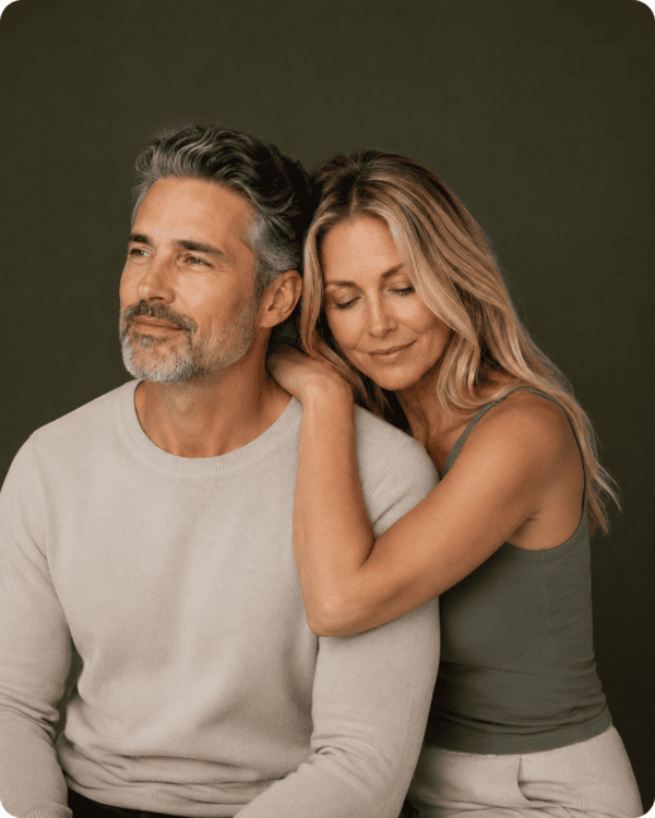 A woman with long blonde hair hugs a bearded man with gray hair from behind. Both appear relaxed and content, sitting close together against a plain dark background.