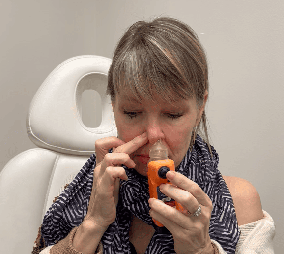A woman sits in a chair using a nasal spray device, holding one nostril closed and her scarf wrapped around her neck.