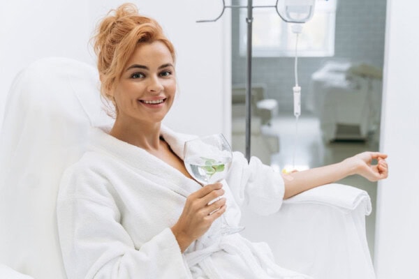 Woman in a white robe smiles while sitting in a clinic chair, holding a glass of water with mint. An IV drip is attached beside her in a bright, clean room.