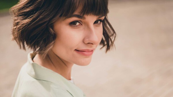 A woman with short brown hair and bangs, wearing a light green top, smiles softly while looking over her shoulder outdoors.